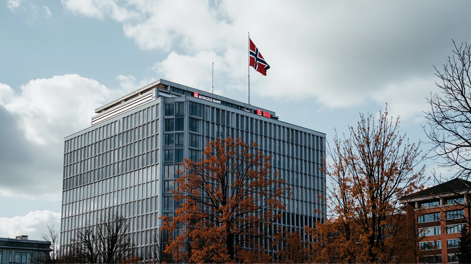 Fachada de la sede del Norges Bank en Oslo bajo cielo otonal nublado, con la bandera noruega ondeando.