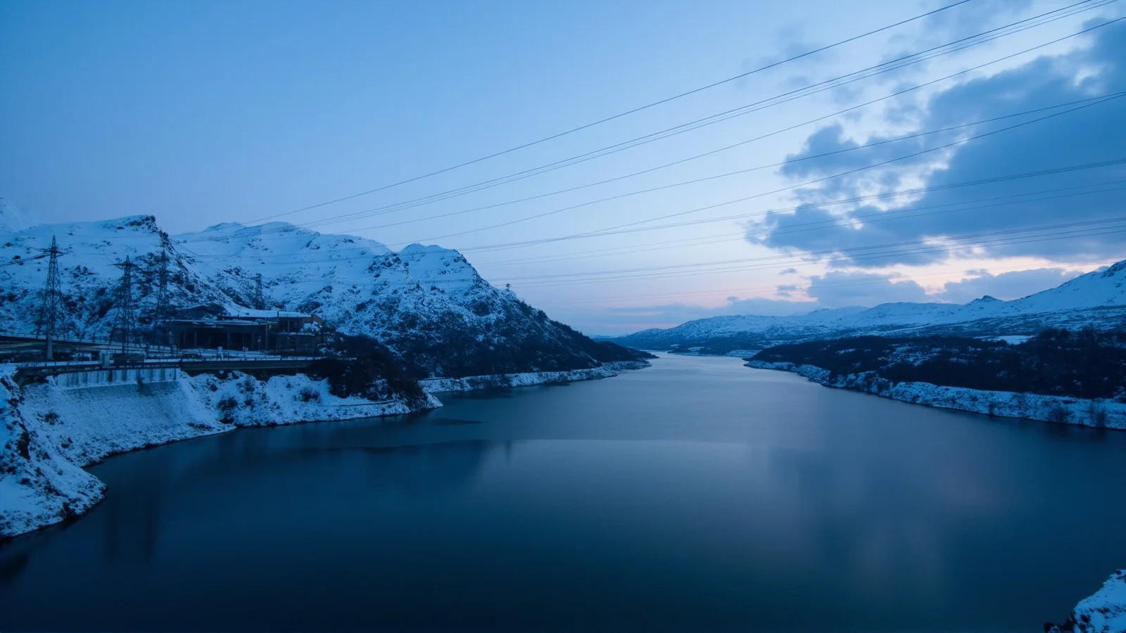 Presa hidroeléctrica noruega al atardecer entre montañas nevadas, con torres de alta tensión sobre el paisaje nevado.